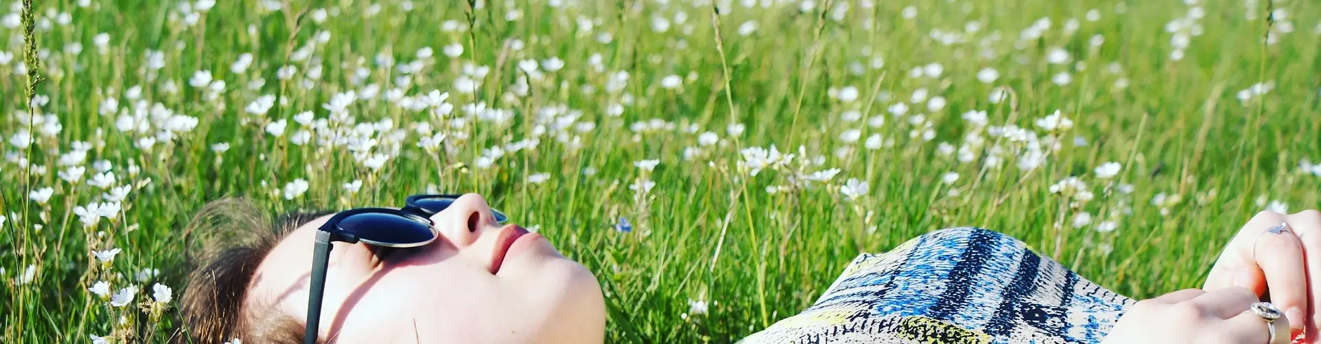 Mujer estirada en un campo de flores blancas y césped verde. Está tranquila y tomando sol. Se podría decir que este campo tiene una firma olfativa ya que el aroma de las flores crea un recuerdo fuerte en esta chica estirada.